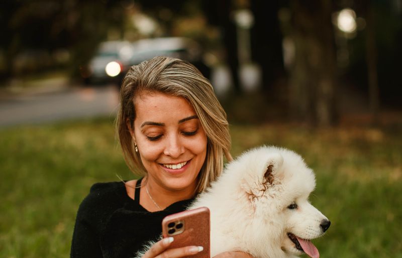 A smiling woman sitting in a park, looking at her phone while holding a fluffy white puppy.